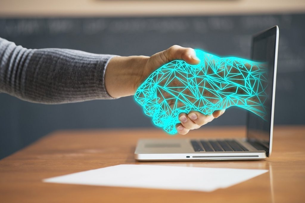 A computerized hand emerges from a laptop to shake hands with a human hand. ChatGPT and customer service in B2B companies.