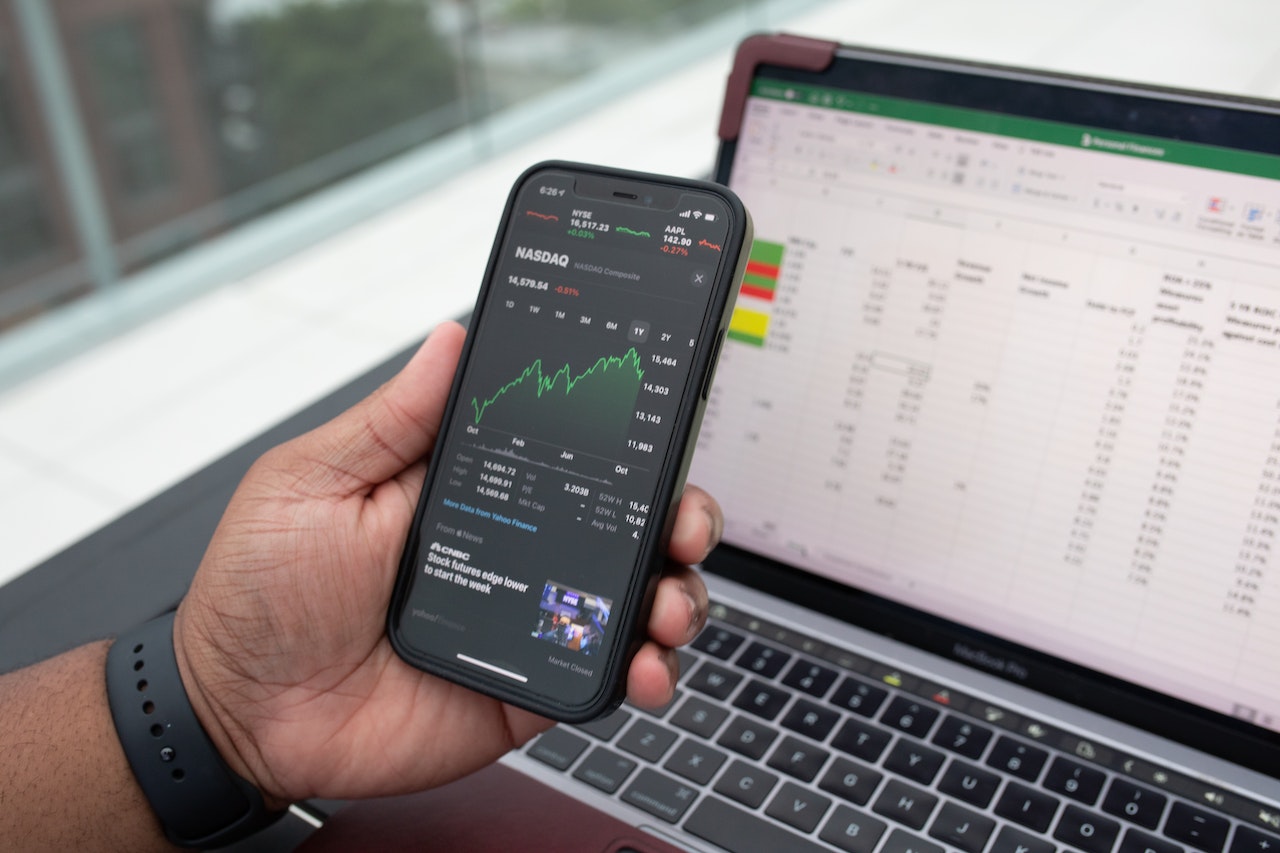 A man's hand holds a phone showing stock information while also viewing a laptop spreadsheet. Client experience in wealth management is turning digital but still must retain a personal touch. 