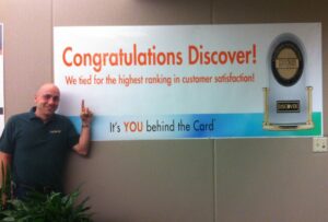 Dan Gingiss poses in front of a banner at Discover headquarters celebrating its first-ever J.D. Power Award win. In recent customer experience news, Capital One announced it is buying Discover Card.