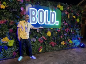 Dan Gingiss stands in front of a colorful flowered wall pointing to a neon sign that reads BOLD. Event experience has become a core focus for planners.